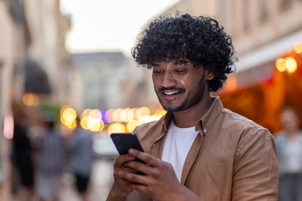 A young Indian man is standing on the street in the city in the evening and using a mobile phone