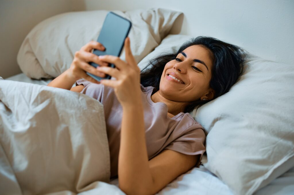 Happy woman texting on mobile phone in bedroom.