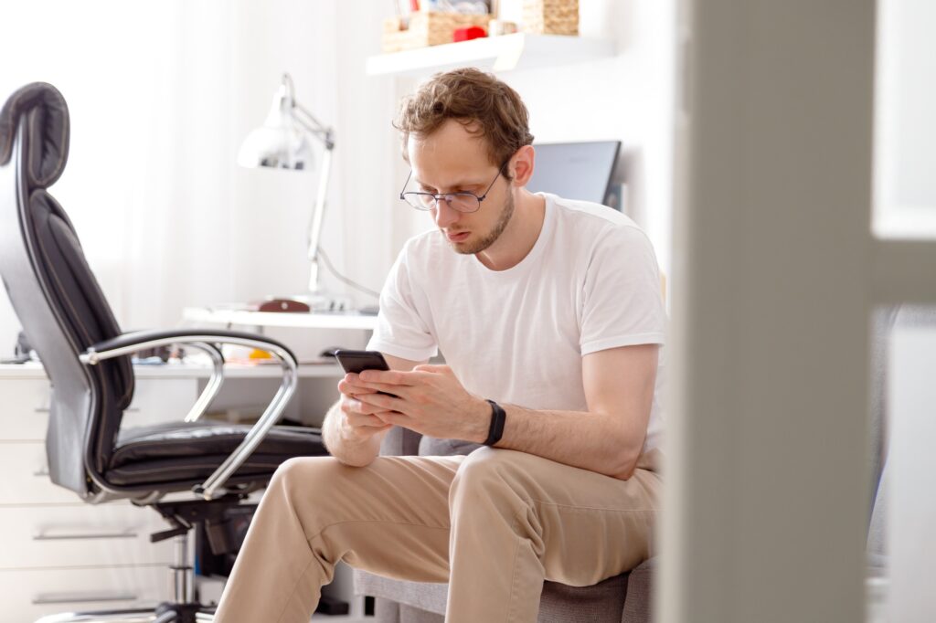 Young man in eyeglasses sitting on sofa sending text on mobile phone